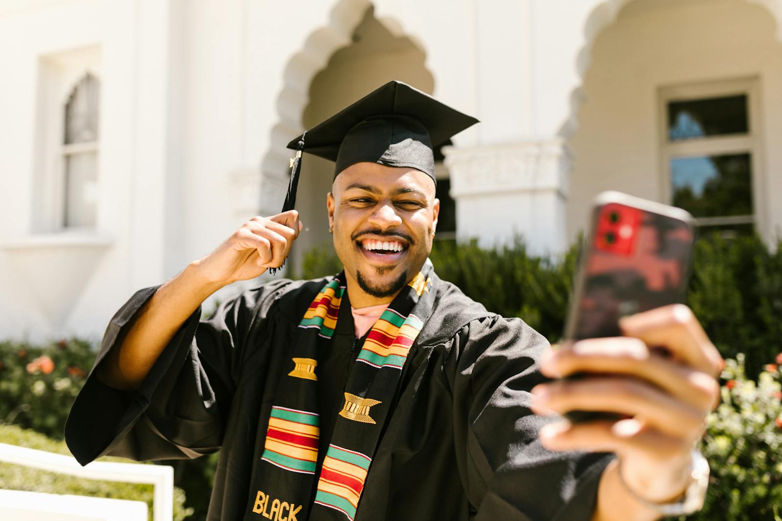 Man in Black Academic Gown Smiling While Holding a Smartphone